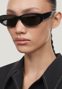 Occhiali da sole neri con montatura elegante e lenti scure, caratterizzati da un design angolare e una finitura lucida, indossati con una camicia nera con colletto.