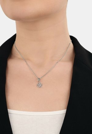 Silberne Halskette mit einem runden Lebensbaum-Anhänger, der ein filigranes Durchbruchdesign aufweist, getragen über einem weißen Hemd.