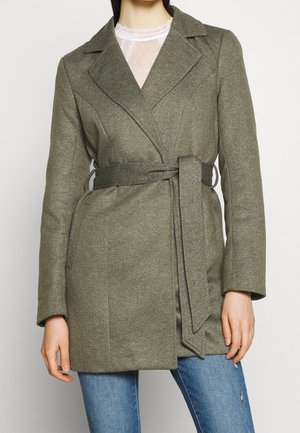 Femme portant un manteau ceinturé vert olive par-dessus un haut en dentelle blanc et un jean bleu, debout devant un fond clair uni.