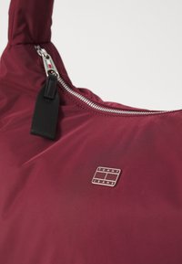 Burgundská taška přes rameno vyrobená z hladkého, odolného materiálu; má výrazný stříbrný zip a nášivku s logem "TOMMY JEANS".