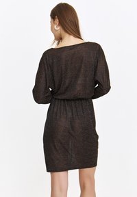 Czarna, dopasowana sukienka z teksturowanej, lśniącej tkaniny. Posiada dekolt typu bateau, długie rękawy i elastyczną talię. Krótka długość.