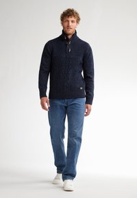 Pulover tricotat în nuanță de bleumarin, cu guler înalt și accente din butoane din lemn, purtat alături de blugi și teniși albi, stând pe un fundal deschis.