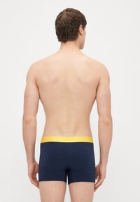 Boxers de algodão azuis com uma cintura elástica amarela com o texto "HAPPY", vistos de costas num torso nu.