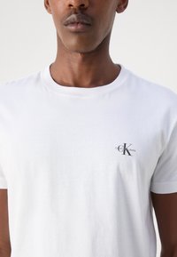 Hvid bomulds t-shirt med rund halsudskæring og et lille sort logo på venstre bryst. Stofserien virker glat og let.
