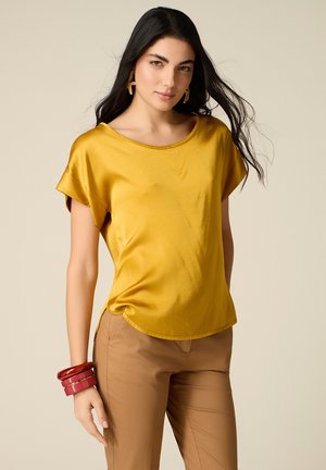 Donna con lunghi capelli scuri che indossa una camicetta di raso giallo senape, pantaloni color cammello, braccialetti rossi e orecchini dorati a forma di stella su sfondo beige.