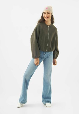 Frau lächelt, trägt eine beige Mütze, eine dunkelgrüne Jacke mit Reißverschluss und Ballonärmeln, hellblaue Schlagjeans und weiße Turnschuhe.