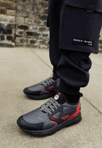 Grå og sorte atletiske sneakers med røde accenter, struktureret overdel og logo-detalje. Har en gummisål og snørebånds lukning.