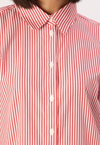 Camicia a righe verticali rossa e bianca con colletto, dotata di chiusura con bottoni. Realizzata in cotone, presenta una texture liscia.