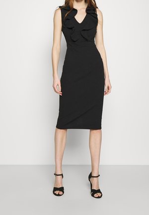 Kobieta w czarnej, bezrękawnej sukience midi z falbanami przy dekolcie w kształcie litery V, w czarnych szpilkach na kostkę, stojąca na tle jednolitego jasnego tła.