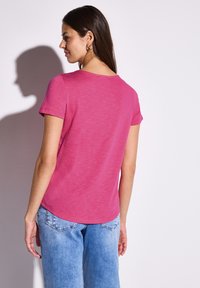 Růžové tričko s krátkým rukávem a kulatým výstřihem, měkká textura a zakřivený lem. Spárováno se světle modrými kalhotami volného střihu.