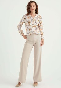 Bloes met bloemenprint in het wit met oranje en paarse accenten, gecombineerd met beige wijde broek en nude puntige hakken.