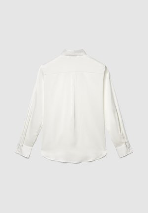 Weiße Bluse mit langen Ärmeln, Manschetten und einer Rückenfalte, flach auf einem hellgrauen Hintergrund liegend dargestellt.