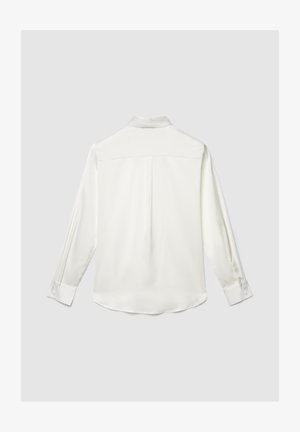 Blusa bianca a maniche lunghe con polsini e piega posteriore, mostrata distesa su uno sfondo grigio chiaro.