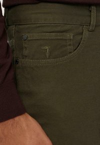 Pantalones verde oliva con un bolsillo frontal, tela texturizada sutil y un pequeño detalle de logo bordado. Presenta detalles con botones metálicos.