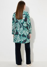 Cappotto lungo a fantasia floreale verde e bianca su uno sfondo scuro, abbinato a pantaloni scuri. Presenta maniche lunghe e una linea dritta.