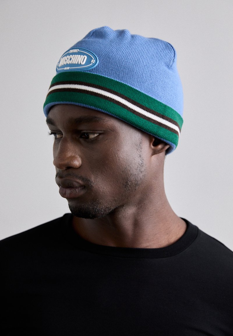 Svjetloplava pletena kapa s plavim i bijelim logom, sa zelenim, smeškim i bijelim prugama na dnu. Glatka tekstura, udobno prianjanje.