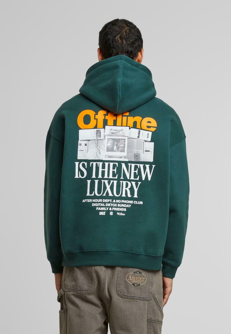 Sudadera verde oscuro con un gran gráfico en texto naranja y blanco en la parte posterior, con un diseño retro de tecnología vintage y texto detallado.