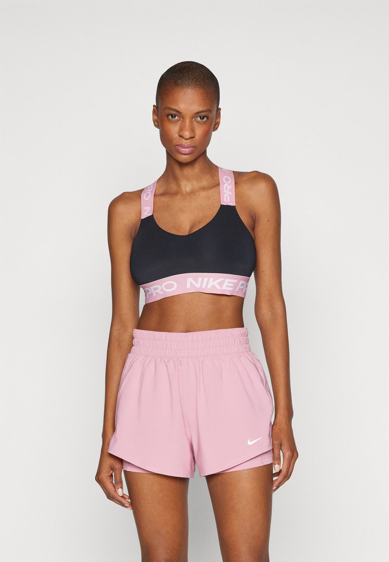 Sostén deportivo negro con tirantes rosa que dicen "PRO NIKE", combinado con pantalones cortos rosa claro que tienen un swoosh blanco de Nike y una cinturilla elástica.