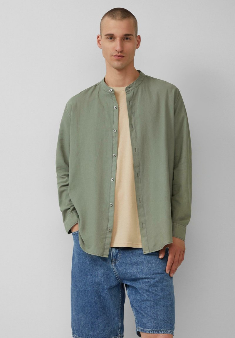 Groene blouse van lichtgewicht stof, met knopen aan de voorkant, gedragen over een beige t-shirt, gecombineerd met denim shorts, staande tegen een neutrale achtergrond.