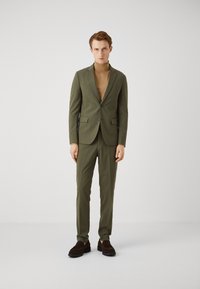 Olive green suit with a slim fit, featuring a single-button blazer, matching trousers, and a brown turtleneck. Suede shoes complete the look.
