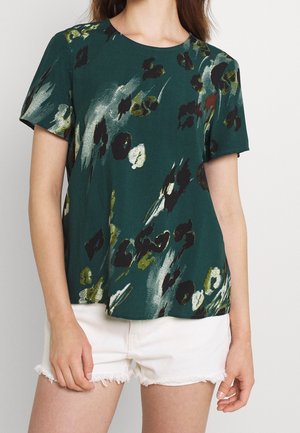 Kobieta ubrana w ciemnozielony top z krótkim rękawem w abstrakcyjny wzór kwiatowy w kolorach czarnym, białym i oliwkowym, w połączeniu z białymi, postrzępionymi szortami jeansowymi.