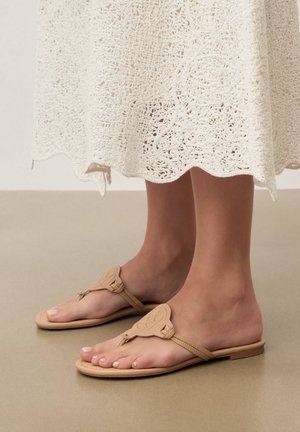 Noge v bež sandalih z vrvico in okrasnim krožnim emblemom, pod belo kvačkano krilo.