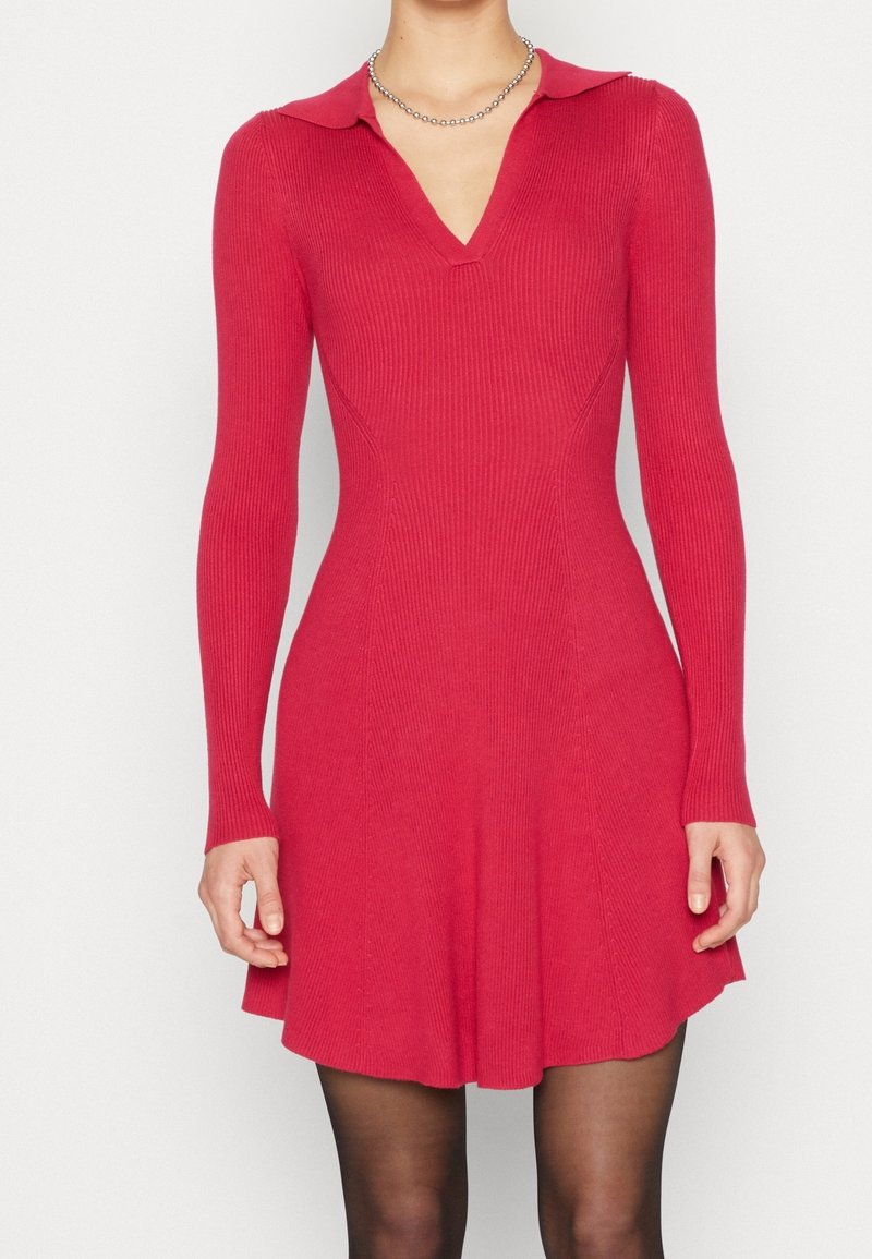 Femme portant une robe en maille rouge cintrée à manches longues avec un col et un décolleté en V, associée à des collants noirs transparents et un collier de perles argenté.