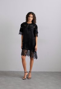 Μαύρο φόρεμα δαντέλας με διαφανές ύφασμα, λουλούδια δαντέλας στα μανίκια και στο τελείωμα, χαλαρή γραμμή, μέχρι το γόνατο, συνδυασμένο με μαύρες ψηλοτάκουνες σανδάλες.