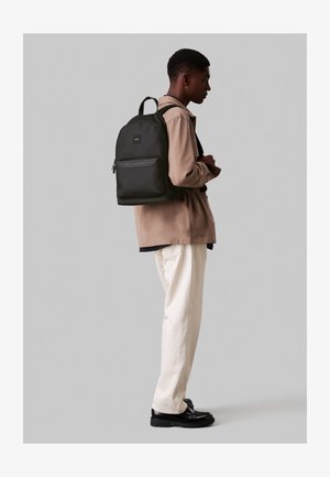 Čierny látkový batoh s prednou vreckom na zips, ergonomickými popruhmi a obdĺžnikovým tvarom, ktorý nosí osoba v oblečení neutrálnych tónov.