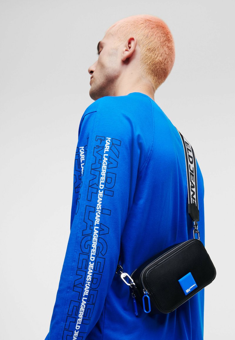 Schwarze Umhängetasche mit blauen Akzenten, rechteckiger Form, mit Reißverschluss und Logodetail, getragen über einem lebhaften blauen Langarmshirt.