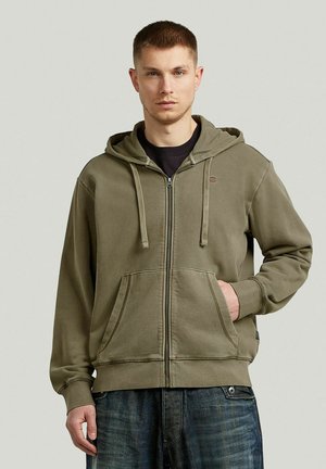 Jeune homme aux cheveux courts portant un sweat à capuche zippé vert olive et un jean bleu, debout avec une main dans la poche.