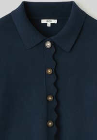 Cardigan boutonné bleu marine avec détail bordé en forme d'ondulation le long de la ligne des boutons et un col côtelé, étiquette taille petite visible.