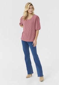 Blusa rosa con occhielli, maniche puff corte e scollatura rotonda, abbinata a jeans blu a campana e mule beige con tacco su sfondo bianco.