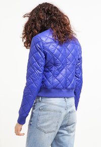 Giacca bomber trapuntata blu con polsini e orlo a coste. Presenta cuciture a forma di rombo e un design cropped, realizzata in materiale sintetico lucido.