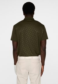 Ciemnooliwkowa koszulka polo z delikatnym geometrycznym wzorem na plecach, krótkimi rękawami i klasycznym kołnierzykiem. Noszona z beżowymi spodniami.