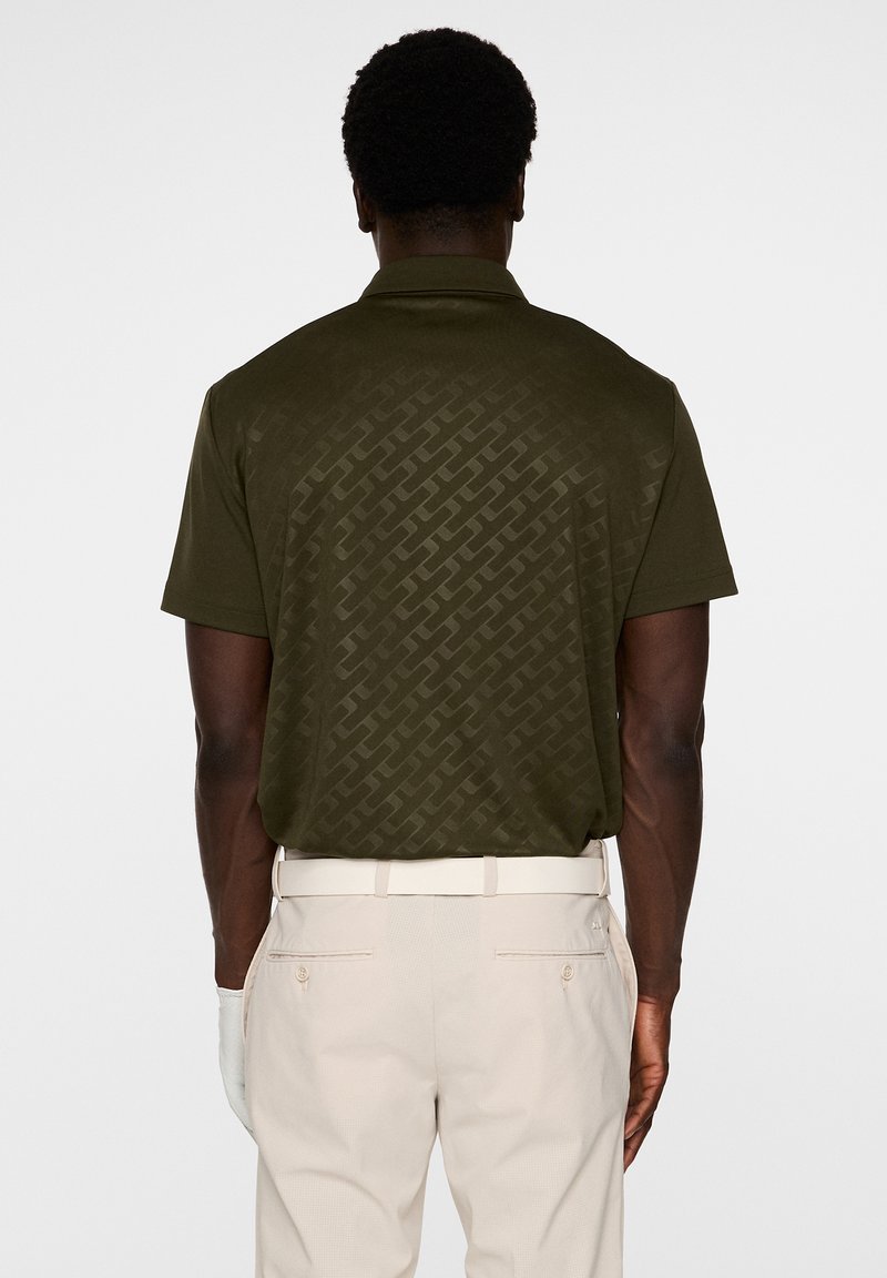 Ciemnooliwkowa koszulka polo z delikatnym geometrycznym wzorem na plecach, krótkimi rękawami i klasycznym kołnierzykiem. Noszona z beżowymi spodniami.