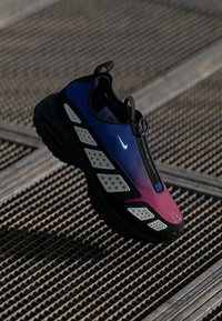 Sneaker sportif avec un dégradé du bleu au rose sur la tige, semelle noire, accents blancs et fermeture éclair sur une surface de grille métallique à motifs.