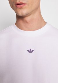 Vit sweatshirt med texturerad yta, prydd med en liten lila broderad logotyp på bröstet. Rund halsringning och ribbade ärmslut.