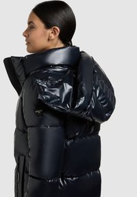 Jachetă puf în material lucios navy, cu guler înalt, logo brodat și buzunare cu fermoar. Textură netedă cu un design modern.