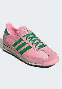 Sapatilha atlética rosa com detalhes verdes, em materiais de camurça e sintéticos, apresentando três riscas, um calcanhar texturizado e atacadores planos.