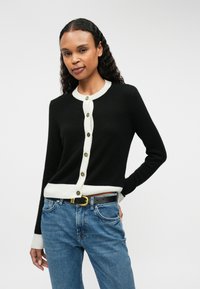 Cardigan noir avec des bordures côtelées crème et des boutons, doté de manches longues. Associé à un jean en denim et une ceinture noire avec une boucle dorée.