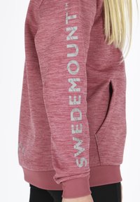 Mörkröd sweatshirt i texturerat tyg, med en logotyptryck i ljusgrått på ärmen och en liten ficka. Ribbstickade mudd och nederkant.