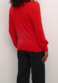 Persoon met krullend haar, gekleed in een rood gestructureerd gebreid trui en zwarte broek, staande tegen een effen lichte muur.