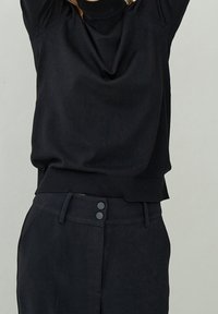 Svart stickad topp med en cowl-nek, ribbad nederkant och avslappnad passform, kombinerad med högmidjade svarta byxor med två framknappar.