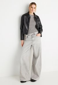 Μαύρο κοντό δερμάτινο τζάκετ πάνω από ριγωτό μακρυμάνικο πουκάμισο, συνδυασμένο με χαλαρά, ανοιχτό γκρι παντελόνια με φαρδιά μπατζάκια και μαύρα παπούτσια.