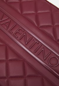 Burgundy läderväska med en quiltad design, präglad "VALENTINO"-logotyp i ett brett band och kontrasterande röda stygn.