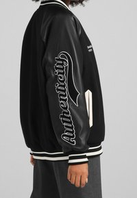 Chaqueta bomber negra con acabado satinado, puños de canalé blancos y "Autenticidad" bordado en la manga; cuenta con bolsillos laterales.