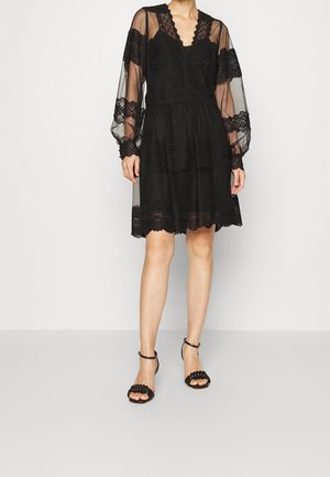 Femme portant une robe noire en dentelle longueur genou avec des manches longues transparentes et des sandales noires à talons hauts et à brides, debout devant un fond uni.