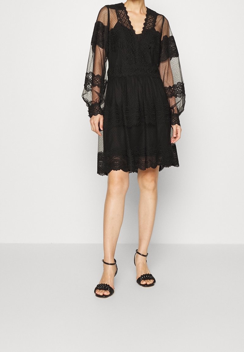 Femme portant une robe noire en dentelle longueur genou avec des manches longues transparentes et des sandales noires à talons hauts et à brides, debout devant un fond uni.
