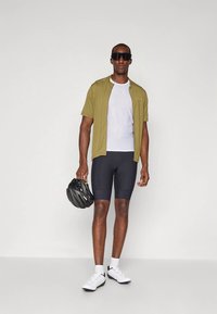 Homme portant un short de cyclisme noir, une chemise en mesh blanche, une veste olive à manches courtes, des chaussures de cyclisme blanches, tenant un casque noir et des lunettes de soleil foncées.